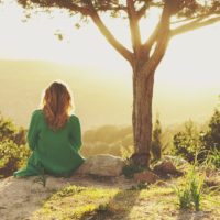 Young woman sitting looking out at sunset on top of a hillside and thinking