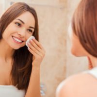 Young woman touching her face with sponge and smiling while standing in front of the mirror
