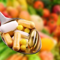 Capsule and pills of vitamins and minerals held in spoon above fruit and vegetables