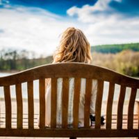 Lonely blonde lady sat on park bench along looking out a lake