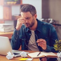 Man in pain sitting by his laptop holding head and squinting