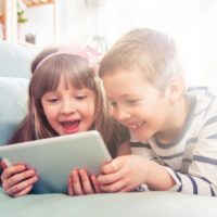 Happy siblings lying on sofa at home and playing with pc tablet together