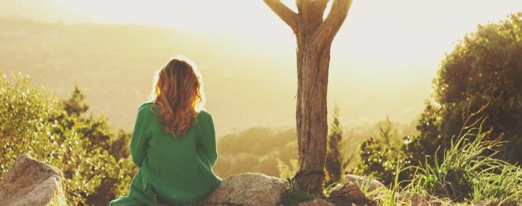 Young woman sitting looking out at sunset on top of a hillside and thinking