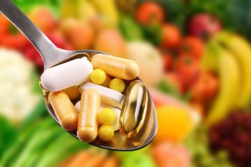 Capsule and pills of vitamins and minerals held in spoon above fruit and vegetables