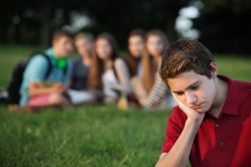 Teenage boy feeling sad being watched by a group of friends in the background