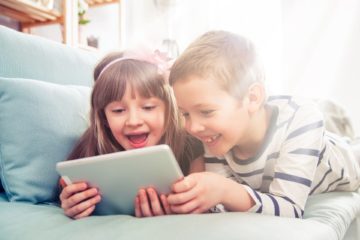 Happy siblings lying on sofa at home and playing with pc tablet together